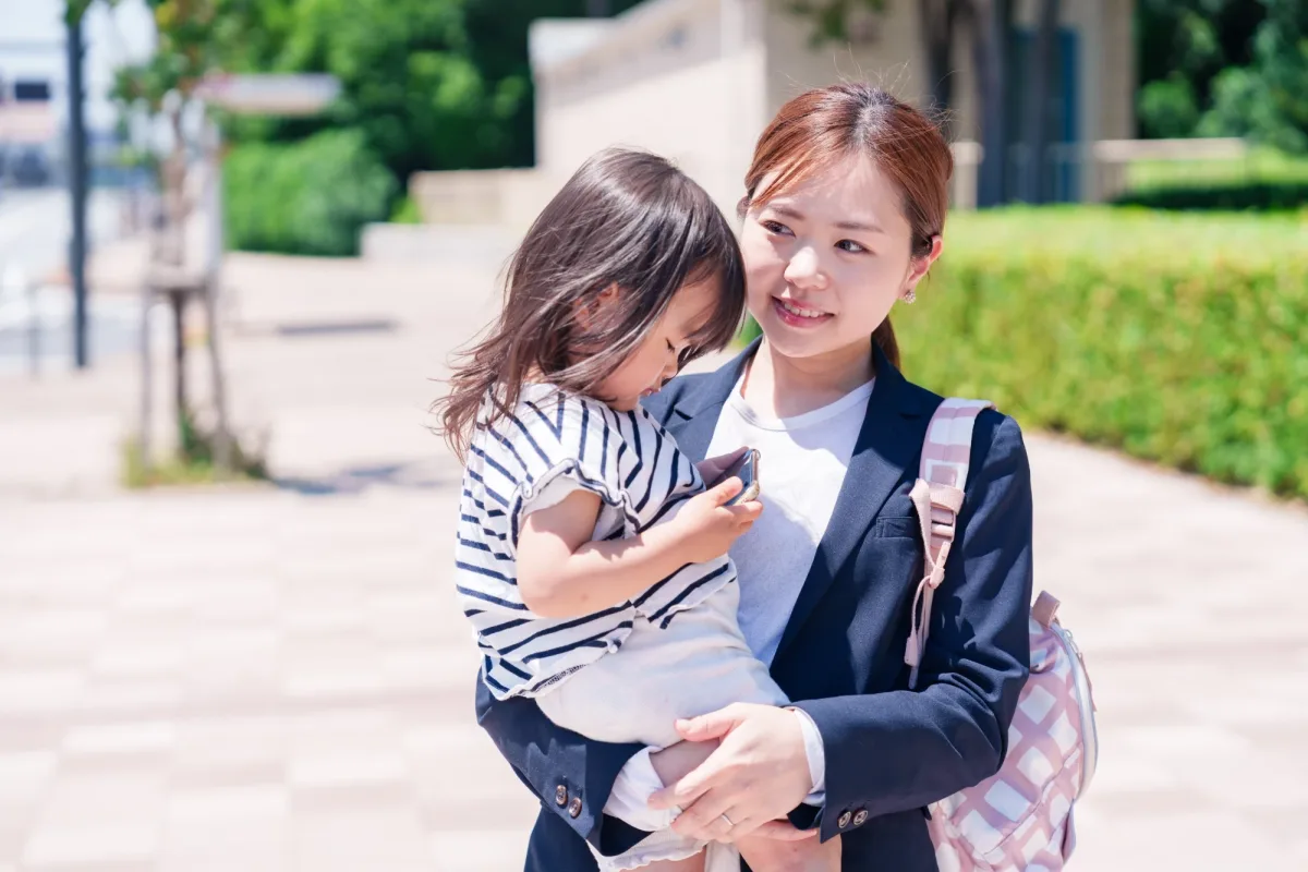 A parent carrying a toddler on a sunny sidewalk